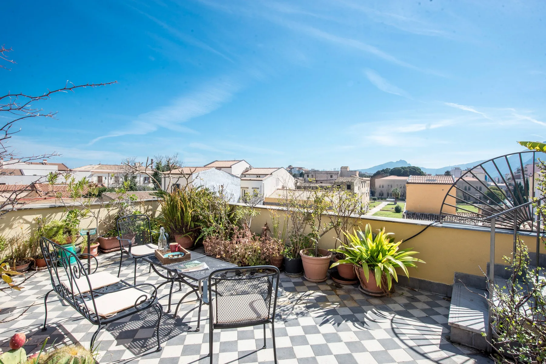 A sunny tiled patio with outdoor seating and potted plants overlooks nearby rooftops.