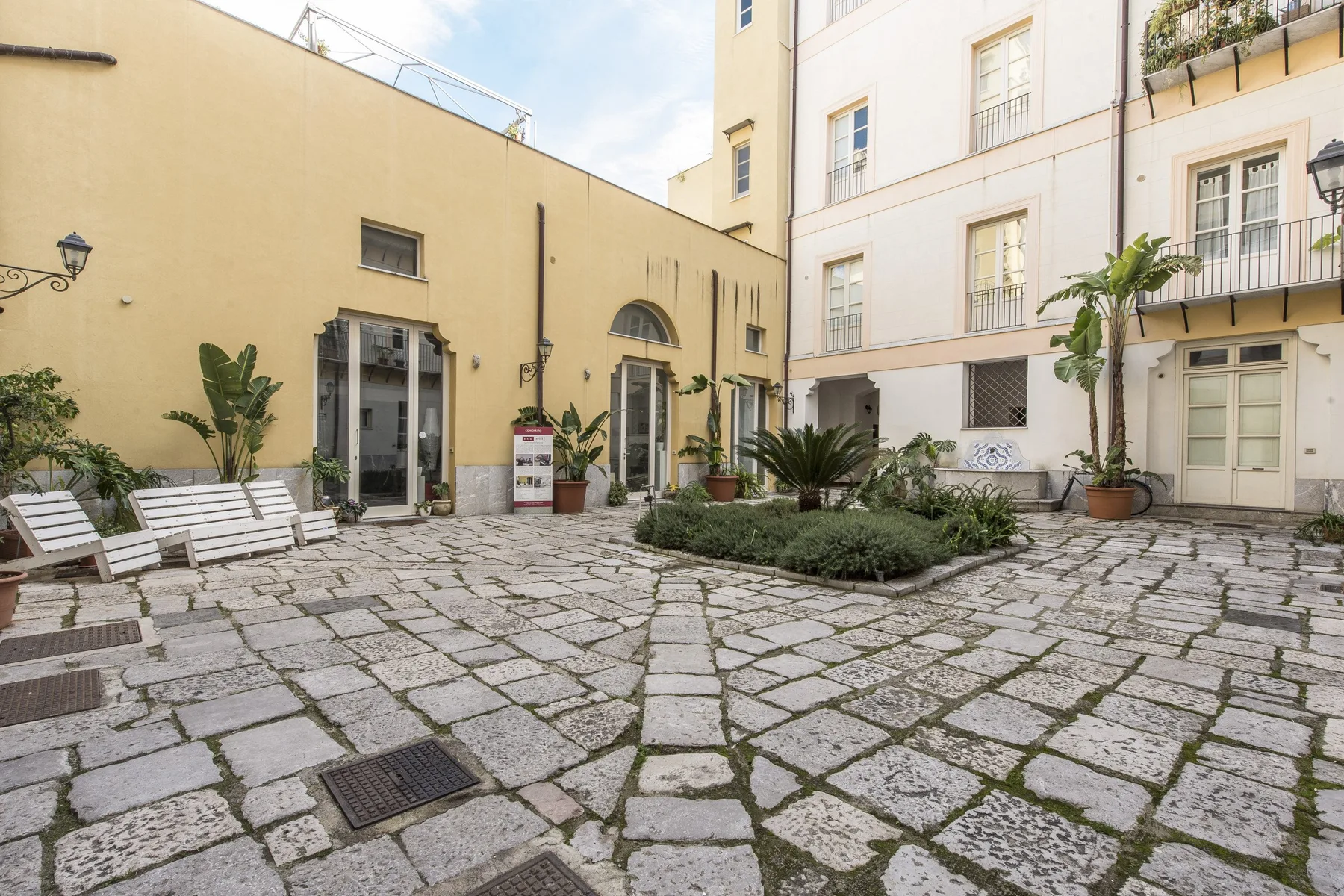 A paved courtyard between light-colored buildings, with a planted garden bed.