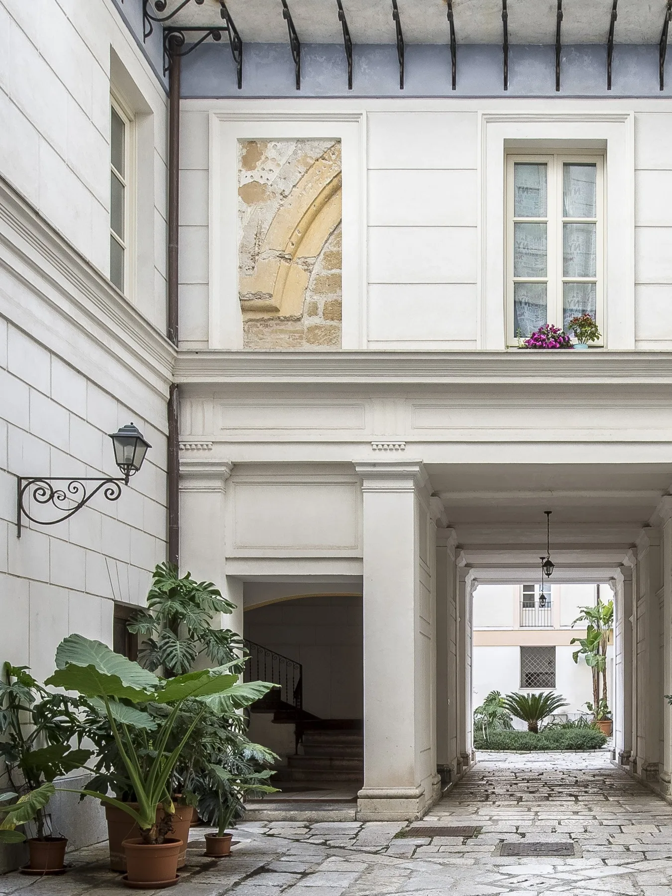A white courtyard passage with stone paving and leafy potted plants.