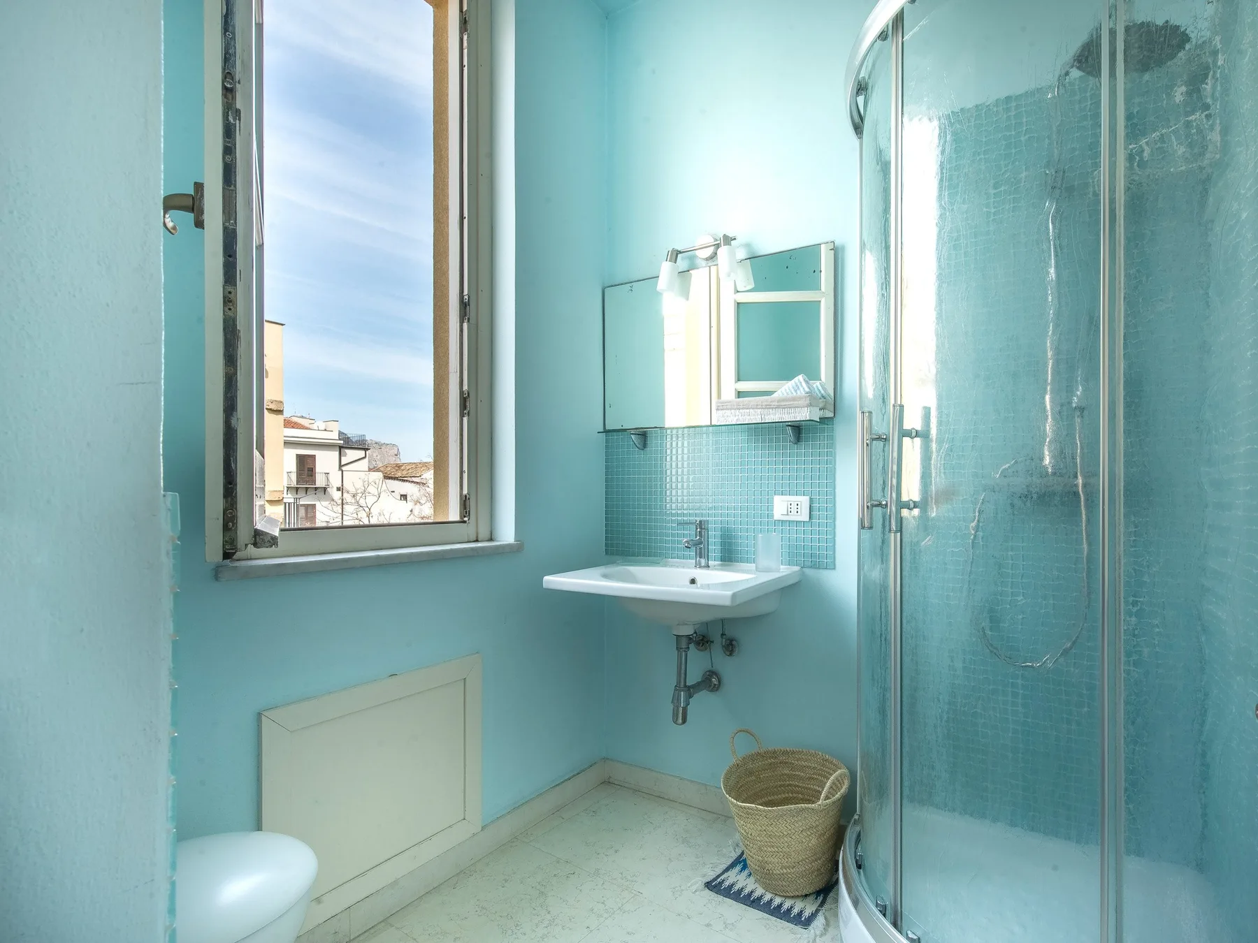 A turquoise bathroom has a sink, mirror, and glass shower beside a window.