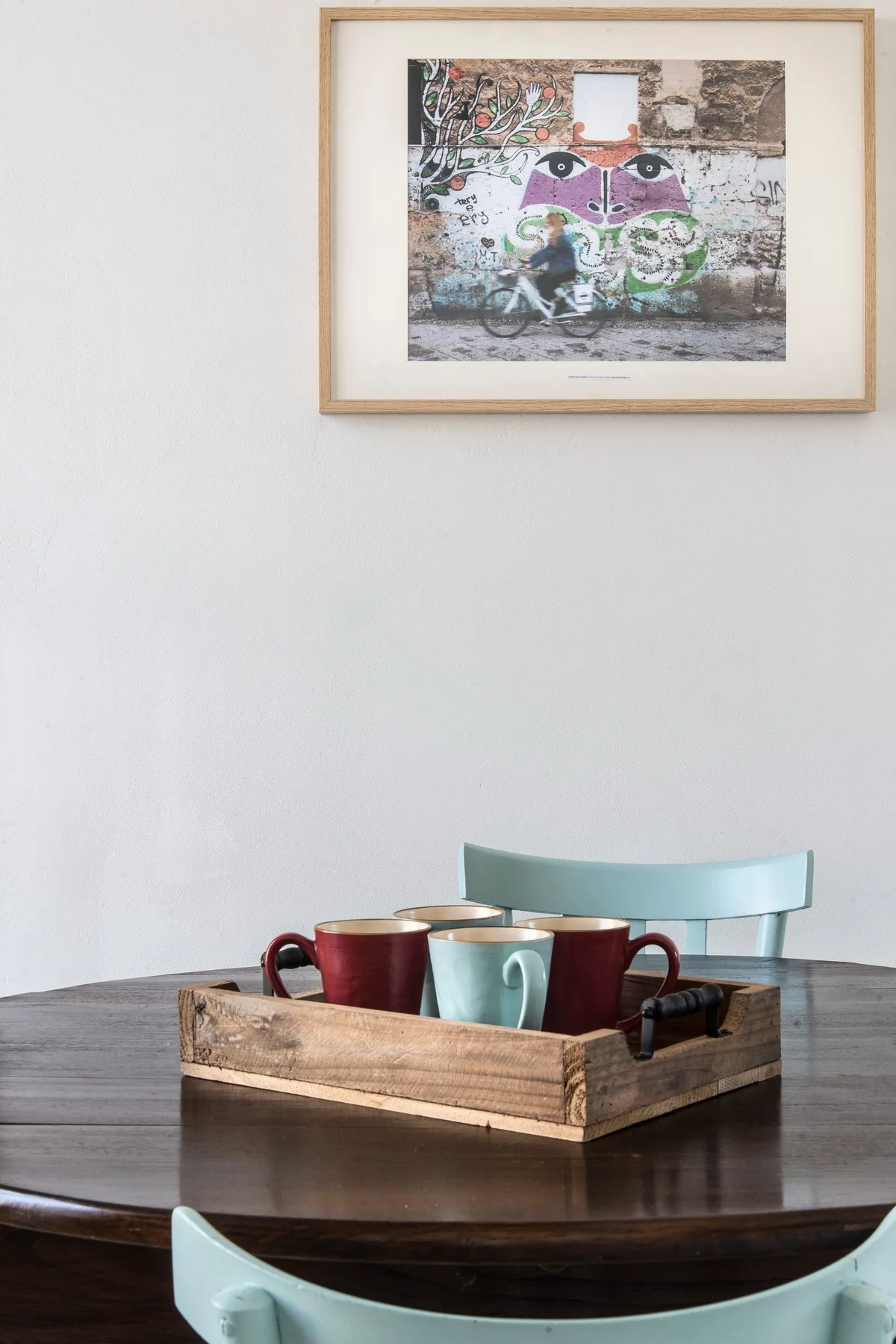 A dining table holds a wooden tray with cups beneath framed wall art.