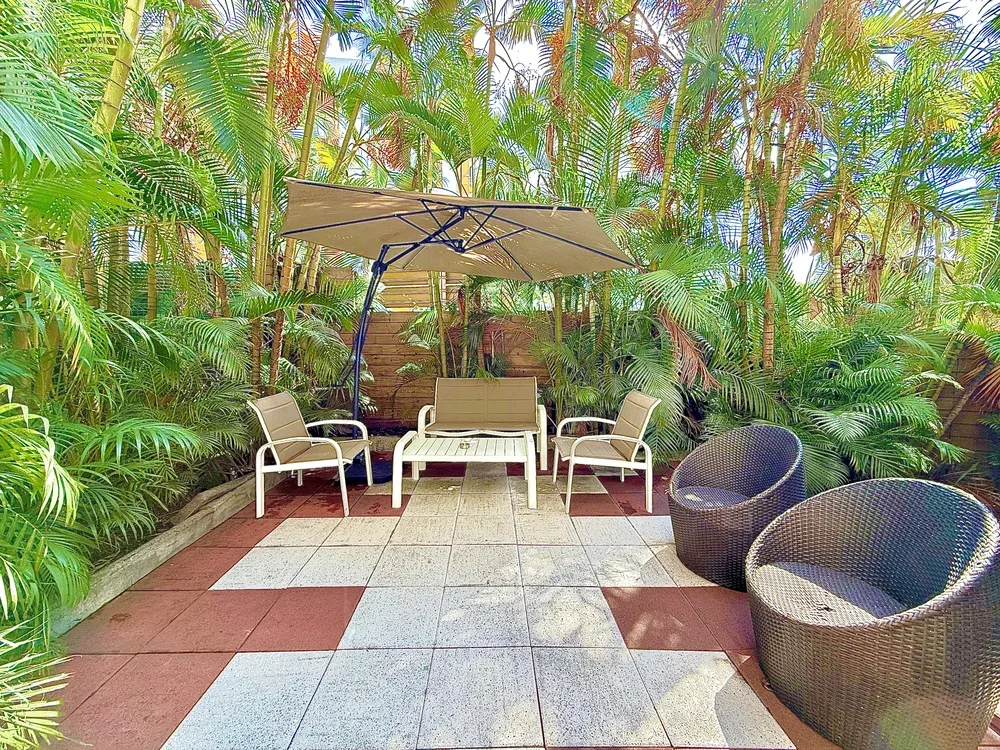 An outdoor patio has seating beneath a large umbrella, surrounded by lush plants.