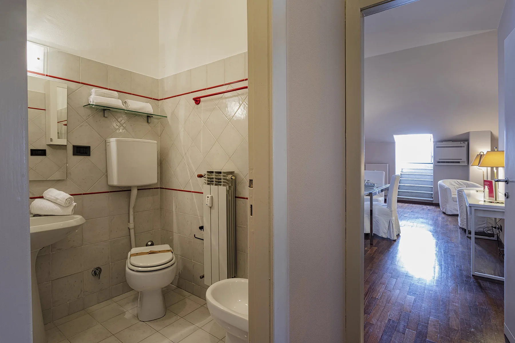 Un baño con inodoro y lavabo da a un dormitorio con camas y un escritorio.