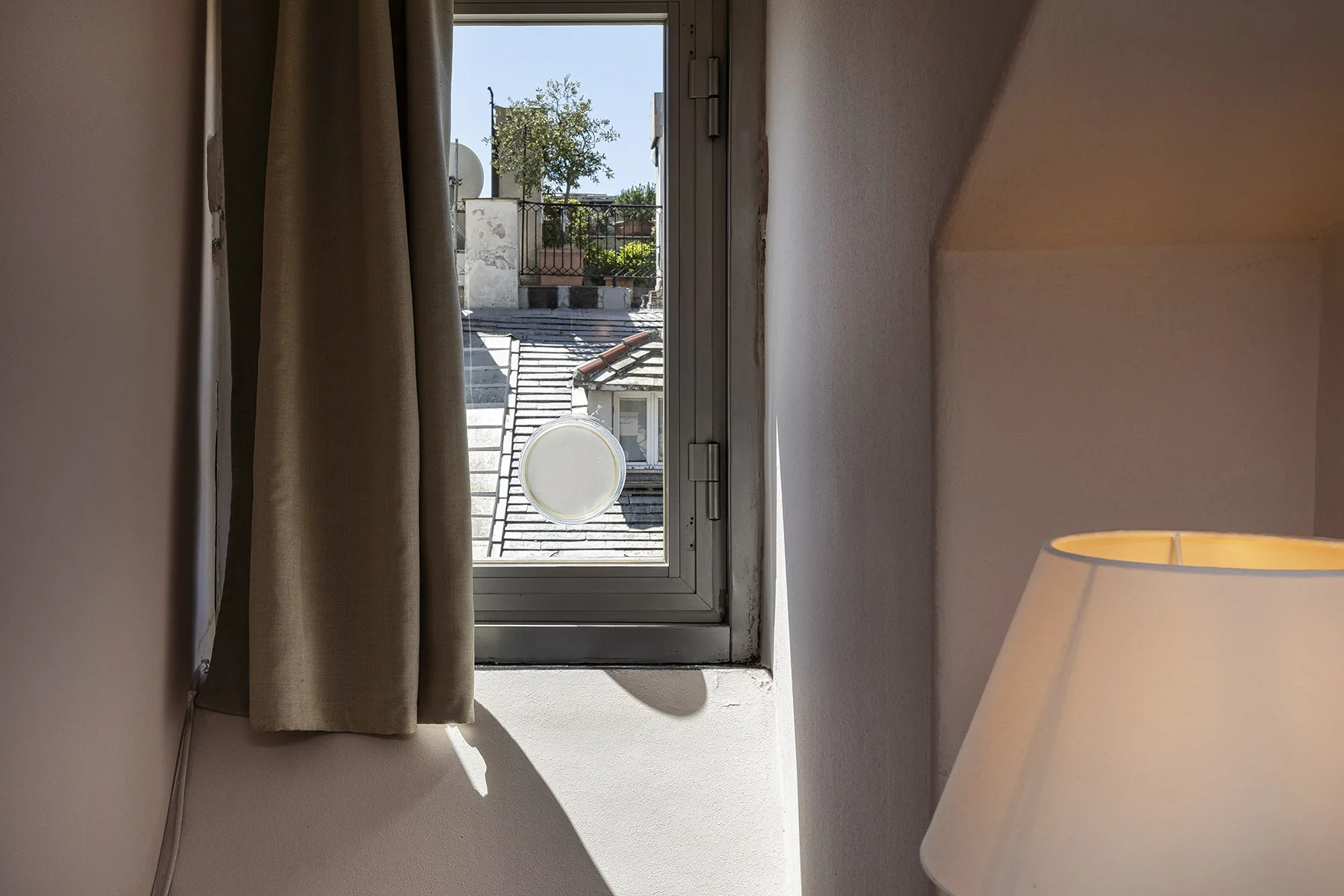 Una ventana con cortina da a una vista de azoteas, con una lámpara encendida cerca.
