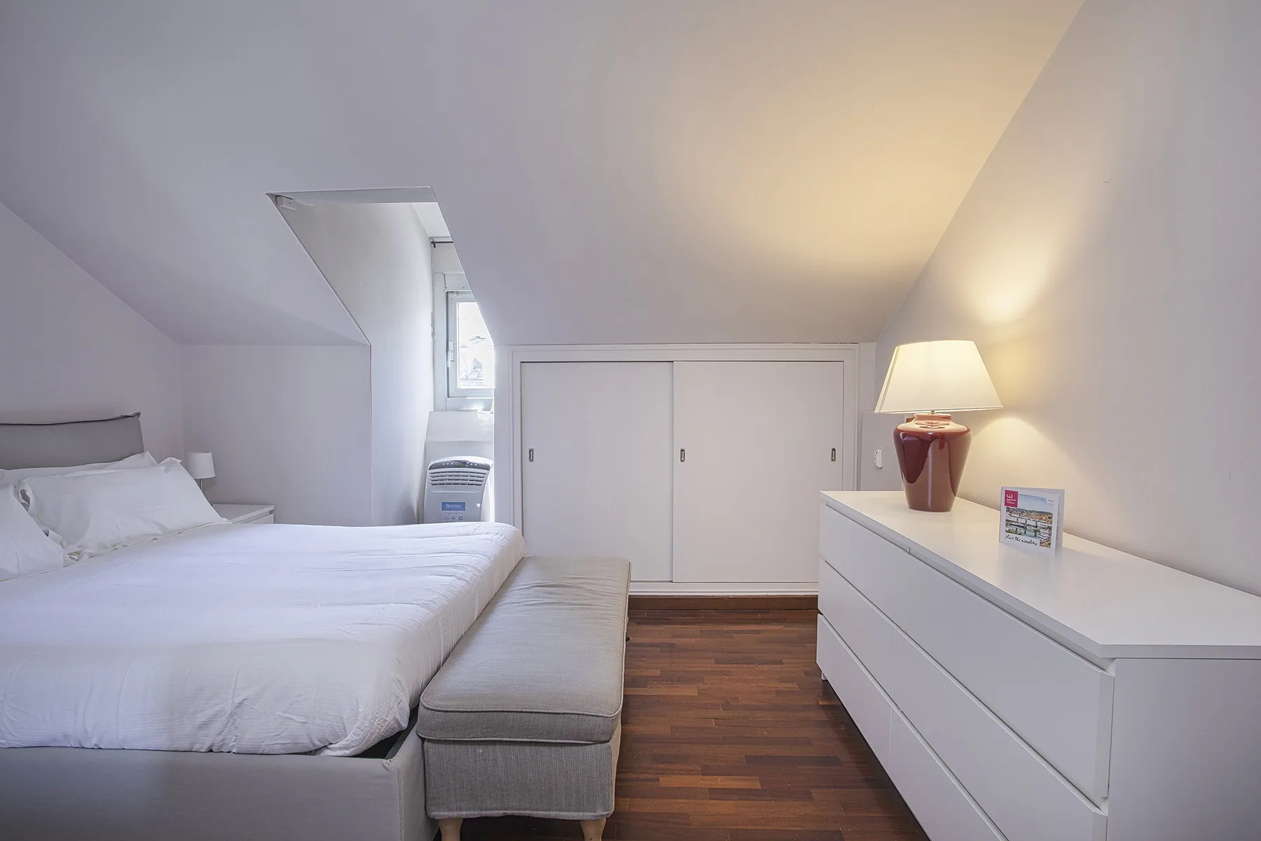 Un dormitorio luminoso con una cama, un banco y una cómoda blanca.