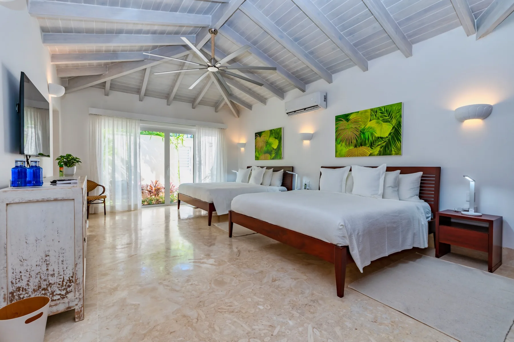 A bright bedroom with two beds, white linens, and green botanical wall art.