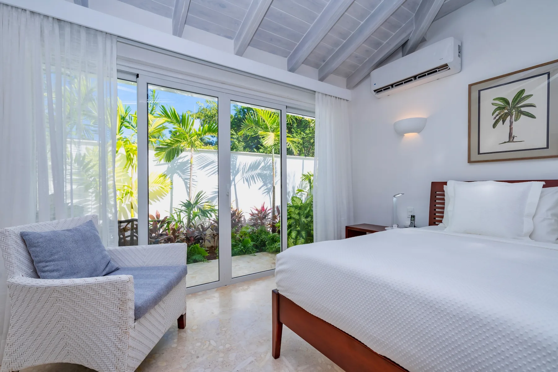 A bright bedroom with a bed, a lounge chair, and glass doors opening to greenery.