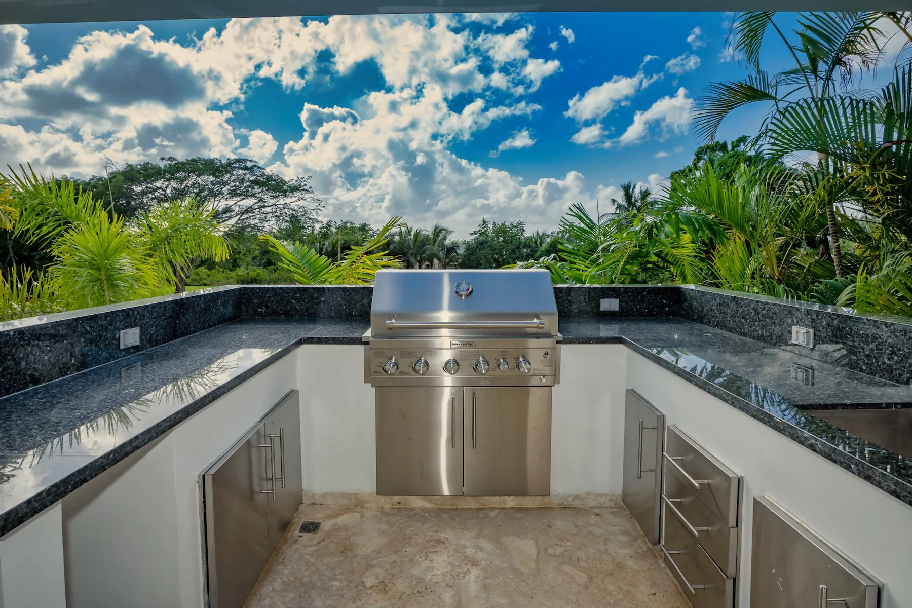 An outdoor kitchen with a built-in grill, surrounded by tropical plants.