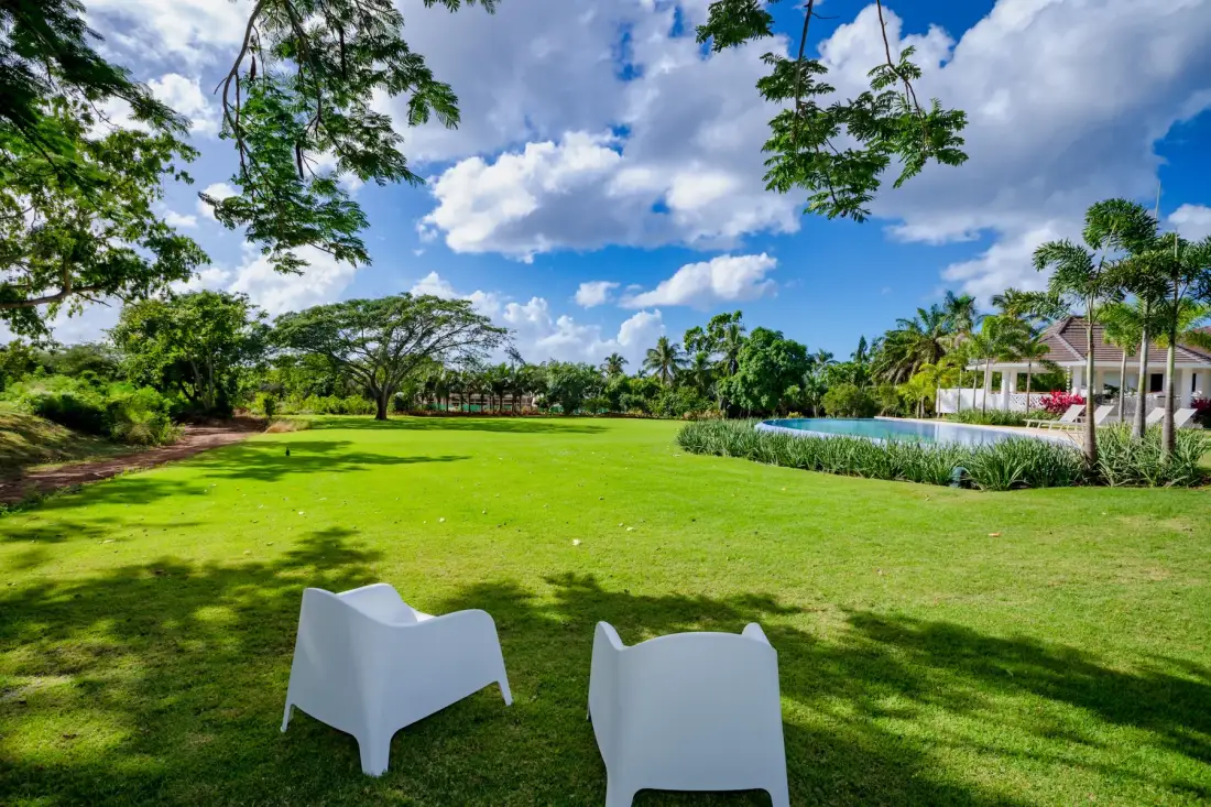 A broad green lawn with two white seats beneath leafy trees.