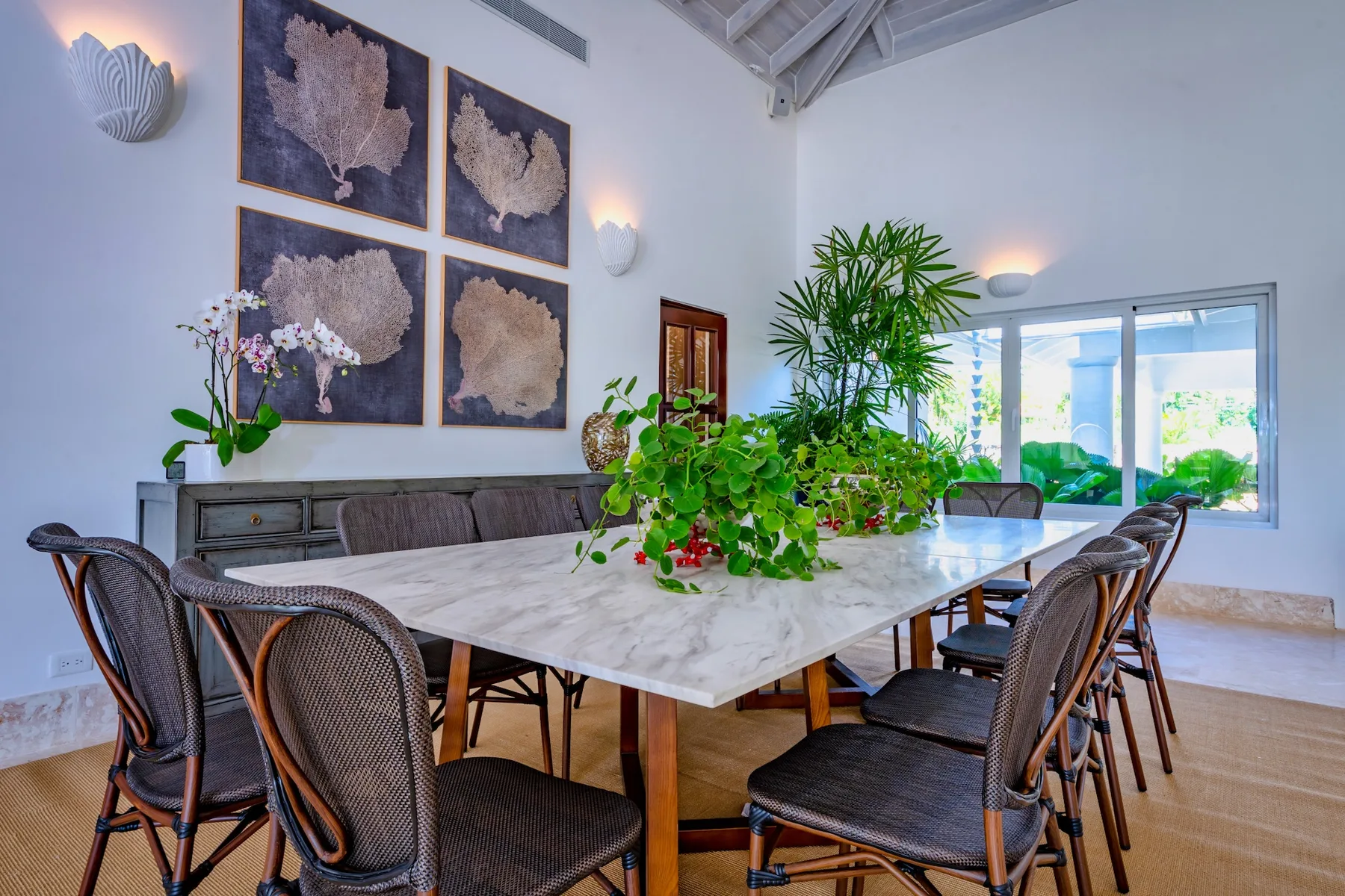 Un comedor luminoso con una mesa larga, asientos tejidos y plantas frondosas.