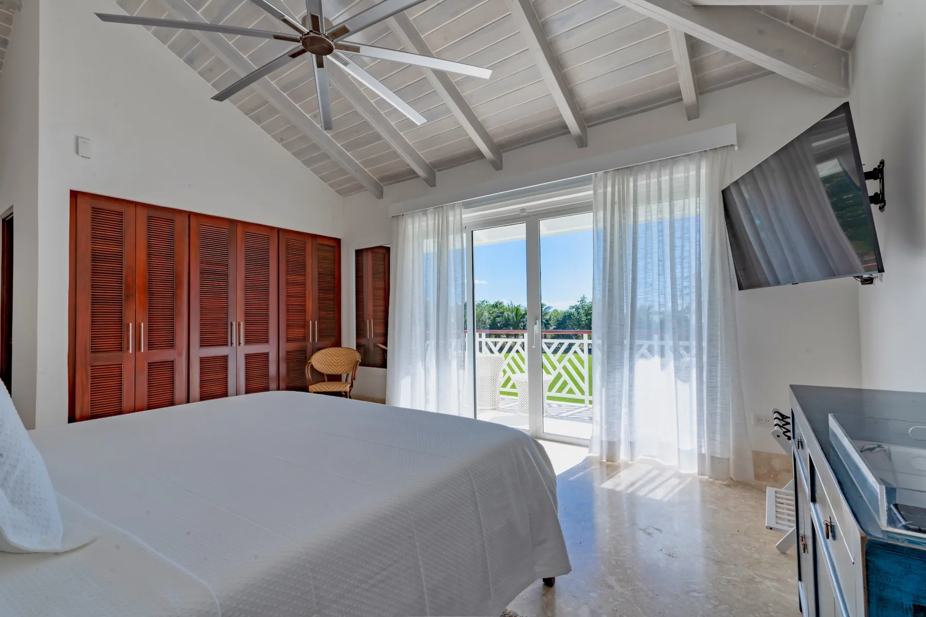 A bright bedroom with a large bed, wooden louvered doors, and a glass door to the balcony.