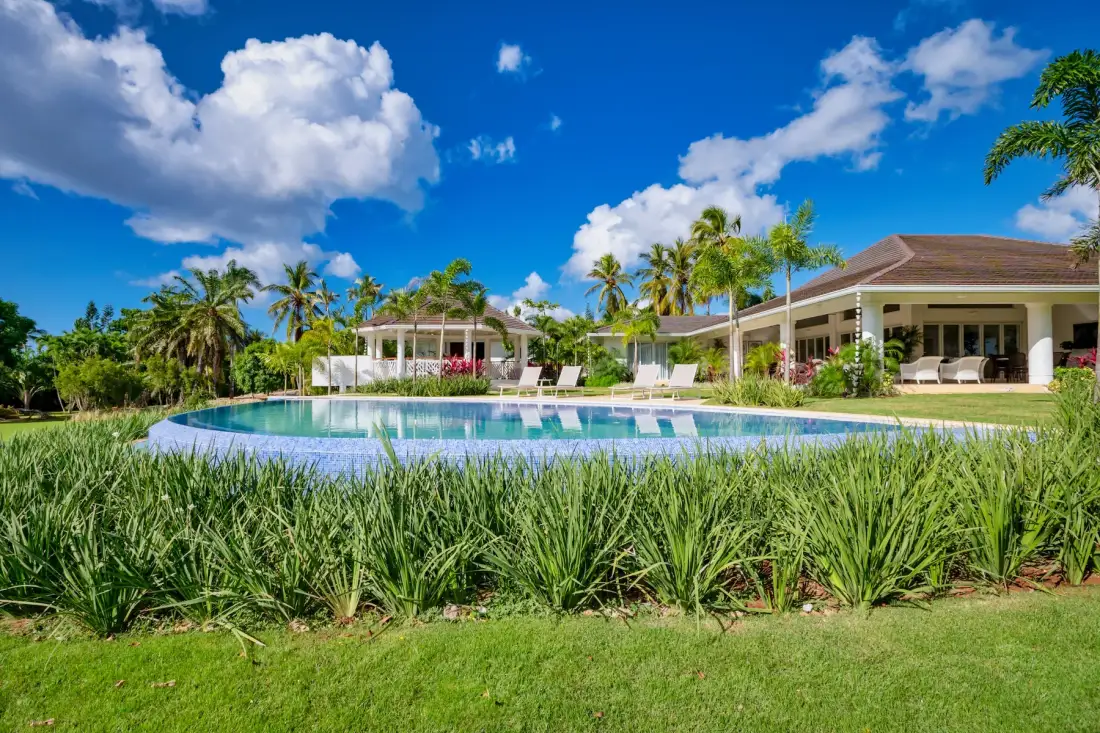 An outdoor pool sits in front of a low resort-style building surrounded by tropical plants.