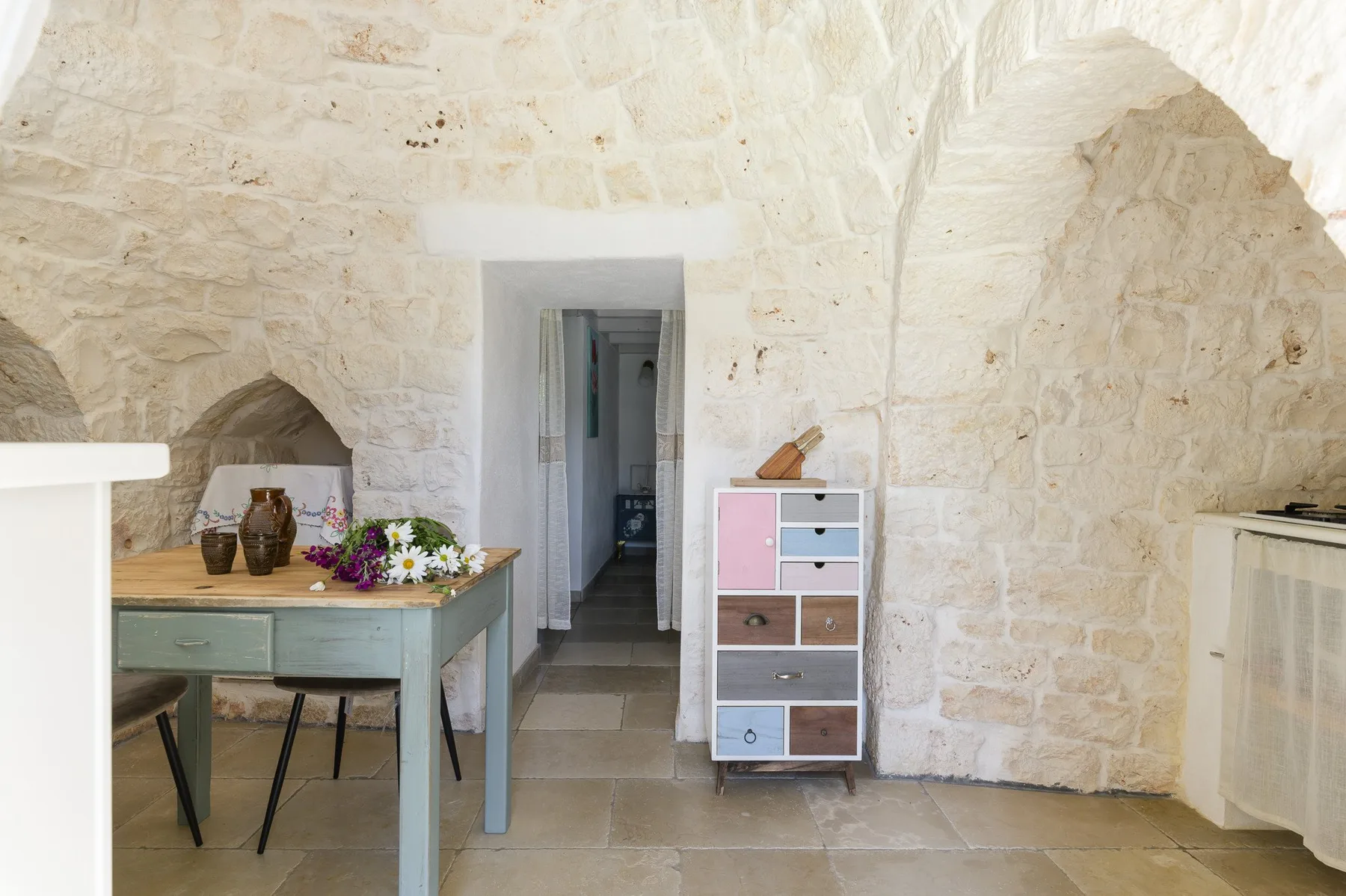 A stone-walled dining area with a table and colorful flowers beside a doorway.
