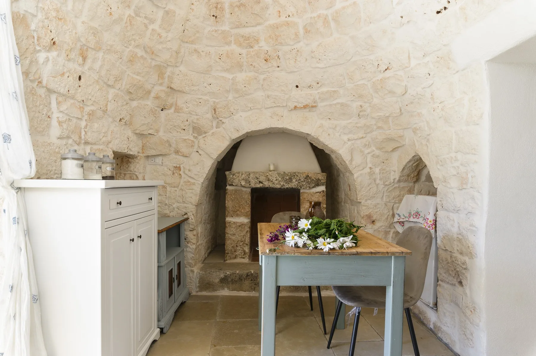 A stone dining area with a wooden table set in front of a fireplace.