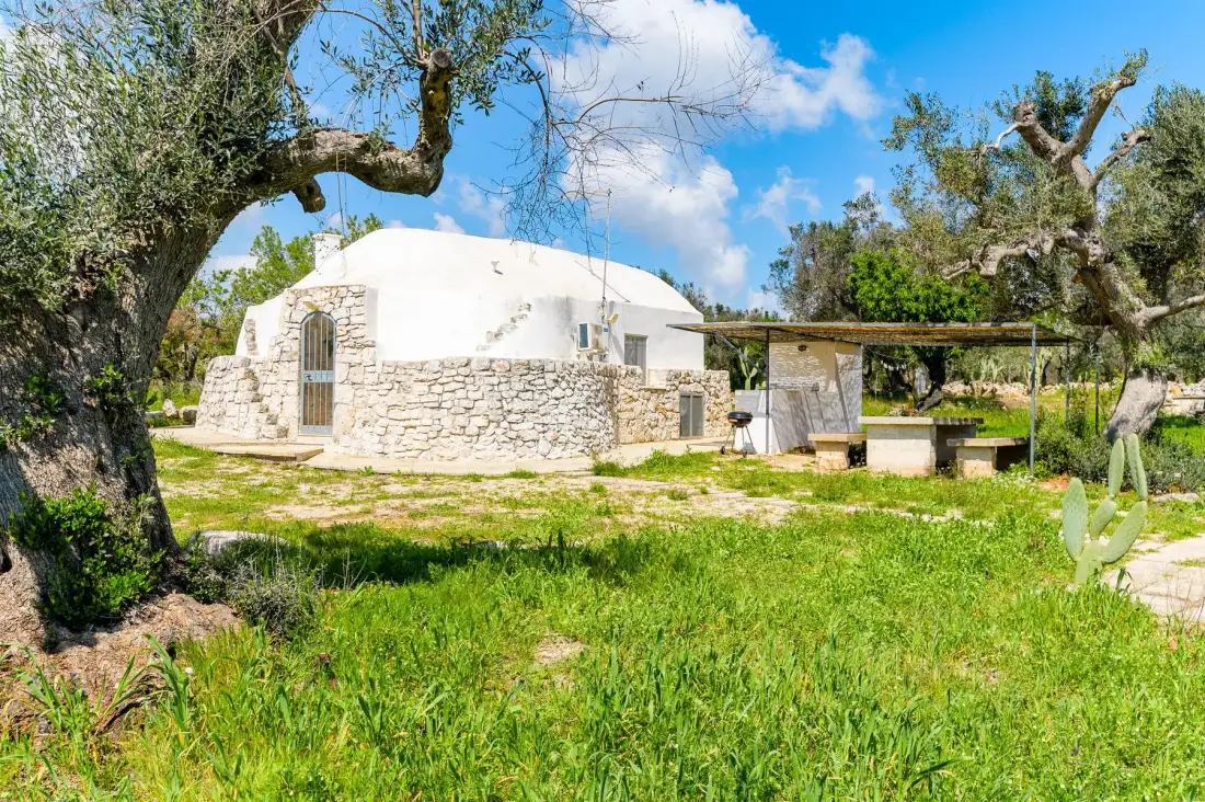 A white stone building stands in a grassy garden beneath mature trees.