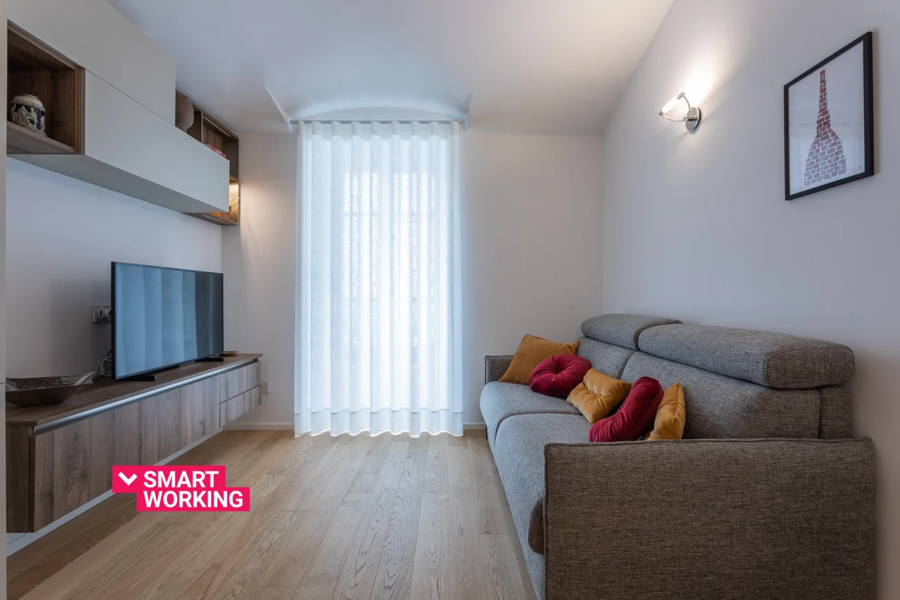 Una sala de estar con televisor, asientos grises y cortinas translúcidas.