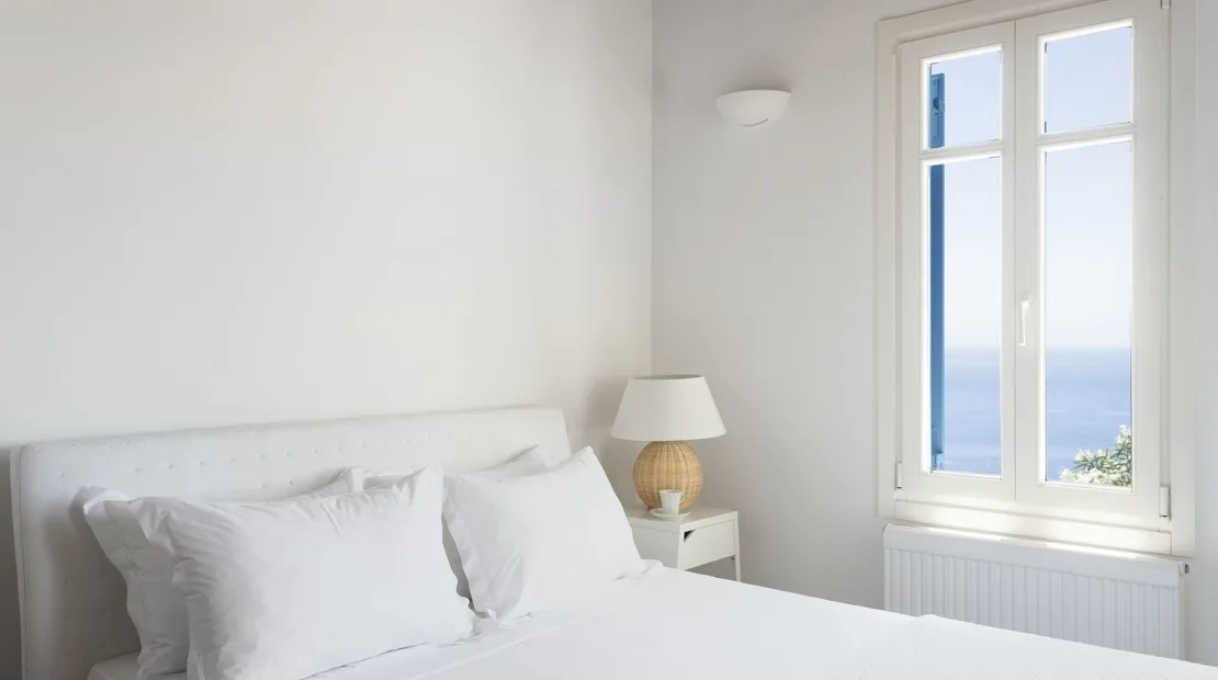 A bright bedroom with white bedding and a window overlooking the sea.