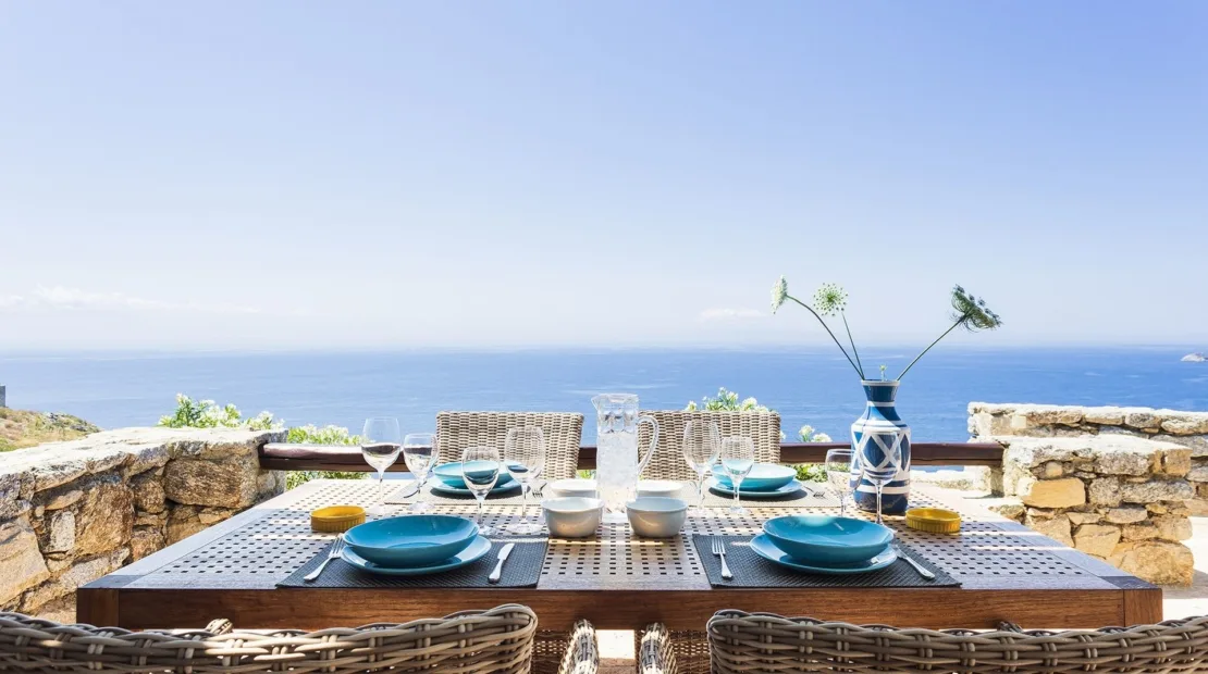 A dining table is set outdoors beside a low stone wall, overlooking the sea.