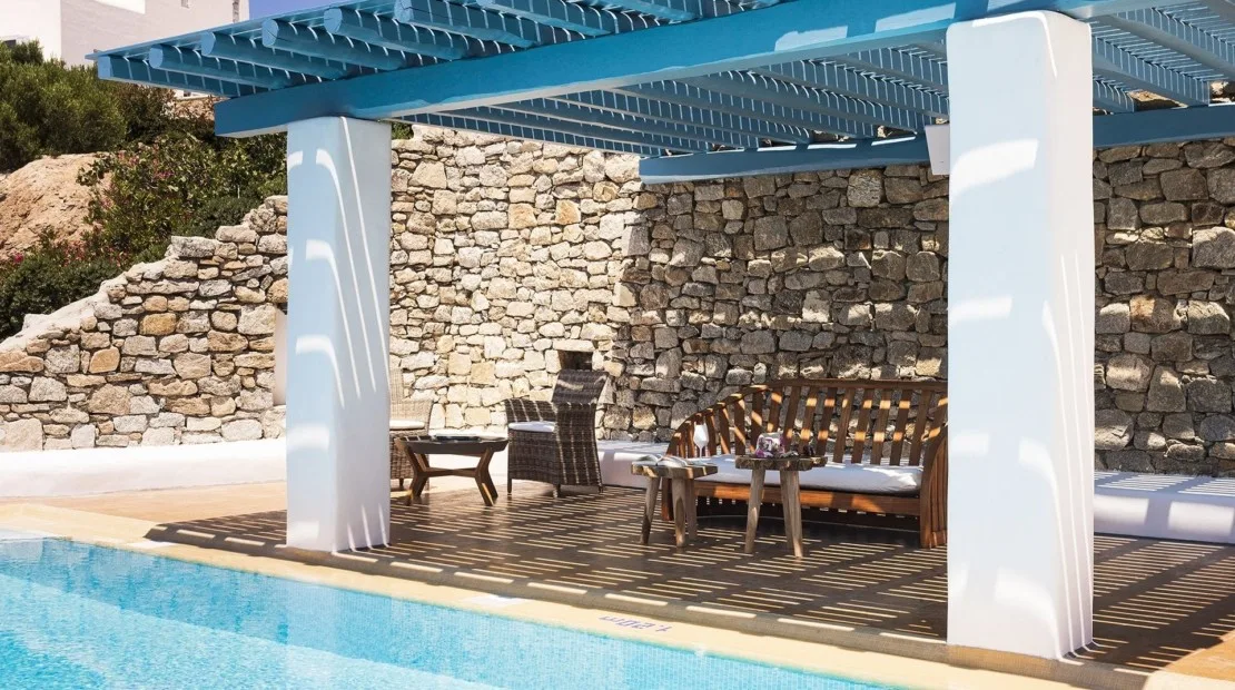 A shaded poolside patio with outdoor seating beneath a blue pergola.