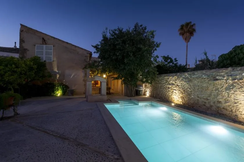 An outdoor swimming pool glows beside a stone wall at dusk.