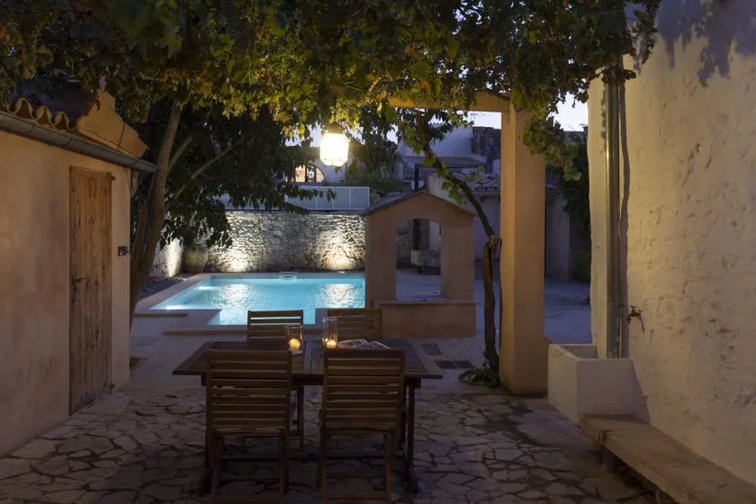 A stone patio with outdoor seating overlooks a lit swimming pool beneath leafy trees.