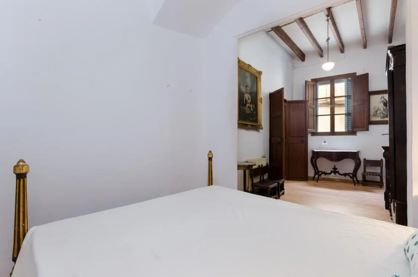 A bedroom with a white bed and an adjoining room with a window and wooden furniture.