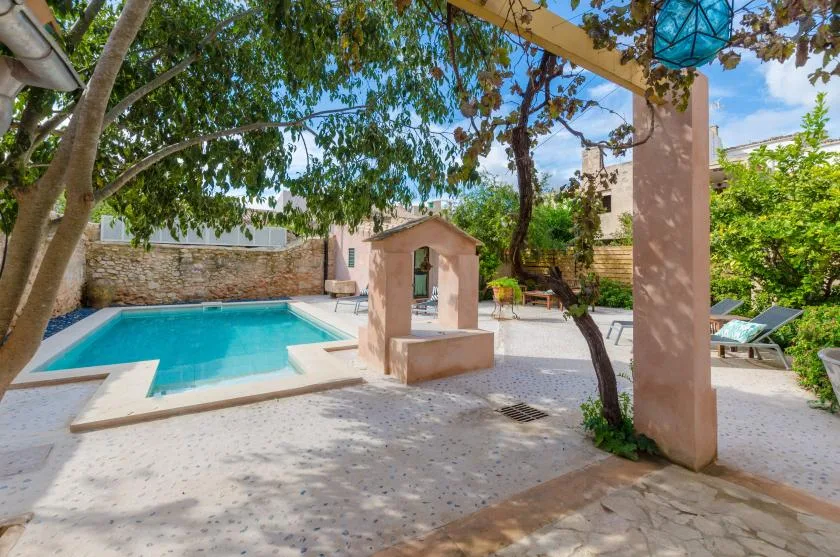 A turquoise swimming pool sits beside a small stone feature in a shaded courtyard.