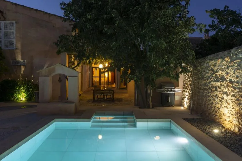 A lit outdoor swimming pool sits beneath trees beside a stone wall.