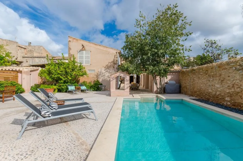 A courtyard swimming pool is surrounded by lounge seating, trees, and stone walls.