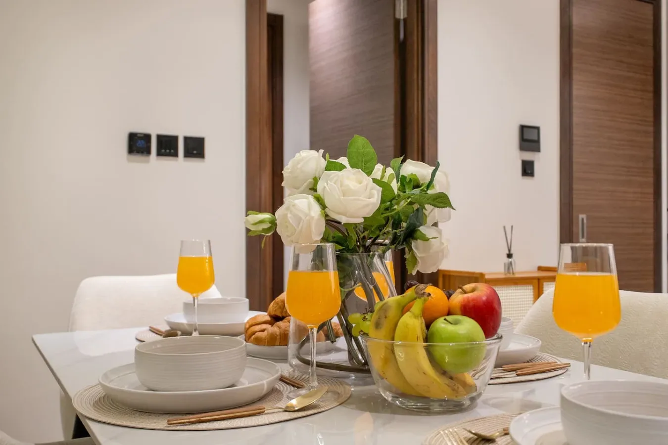 Una mesa de comedor está puesta con platos, fruta, flores y bebidas anaranjadas.