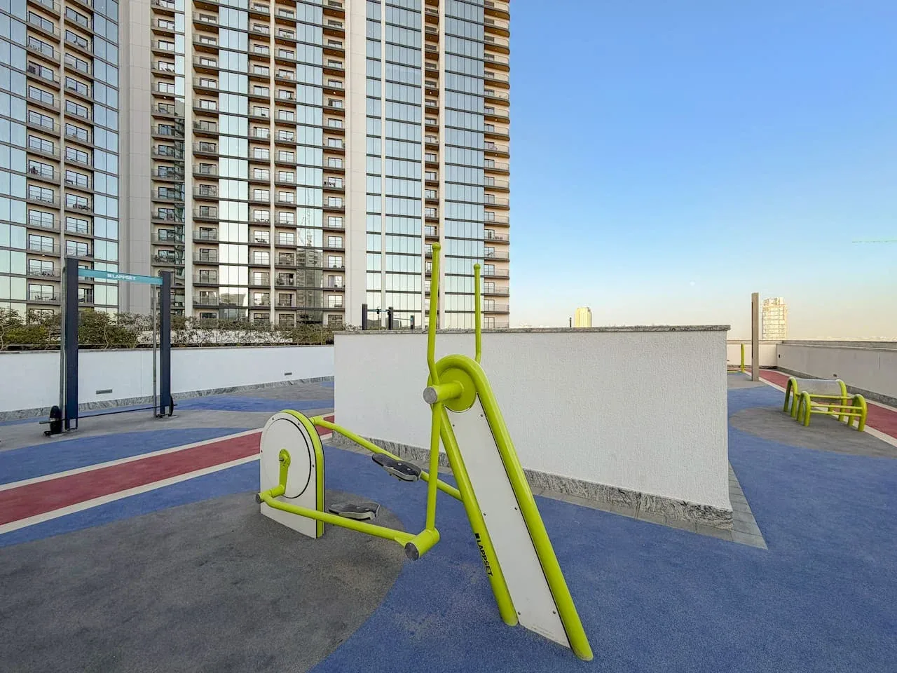Una zona de ejercicio al aire libre con aparatos verdes y edificios altos al fondo.
