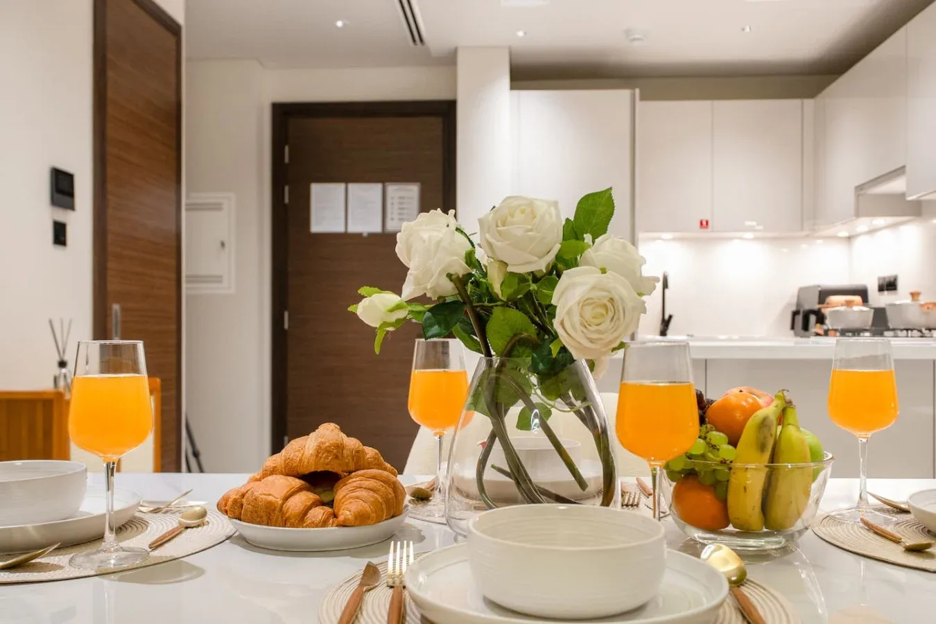 Una mesa puesta con zumo de naranja, bollería, fruta y flores blancas en una cocina luminosa.
