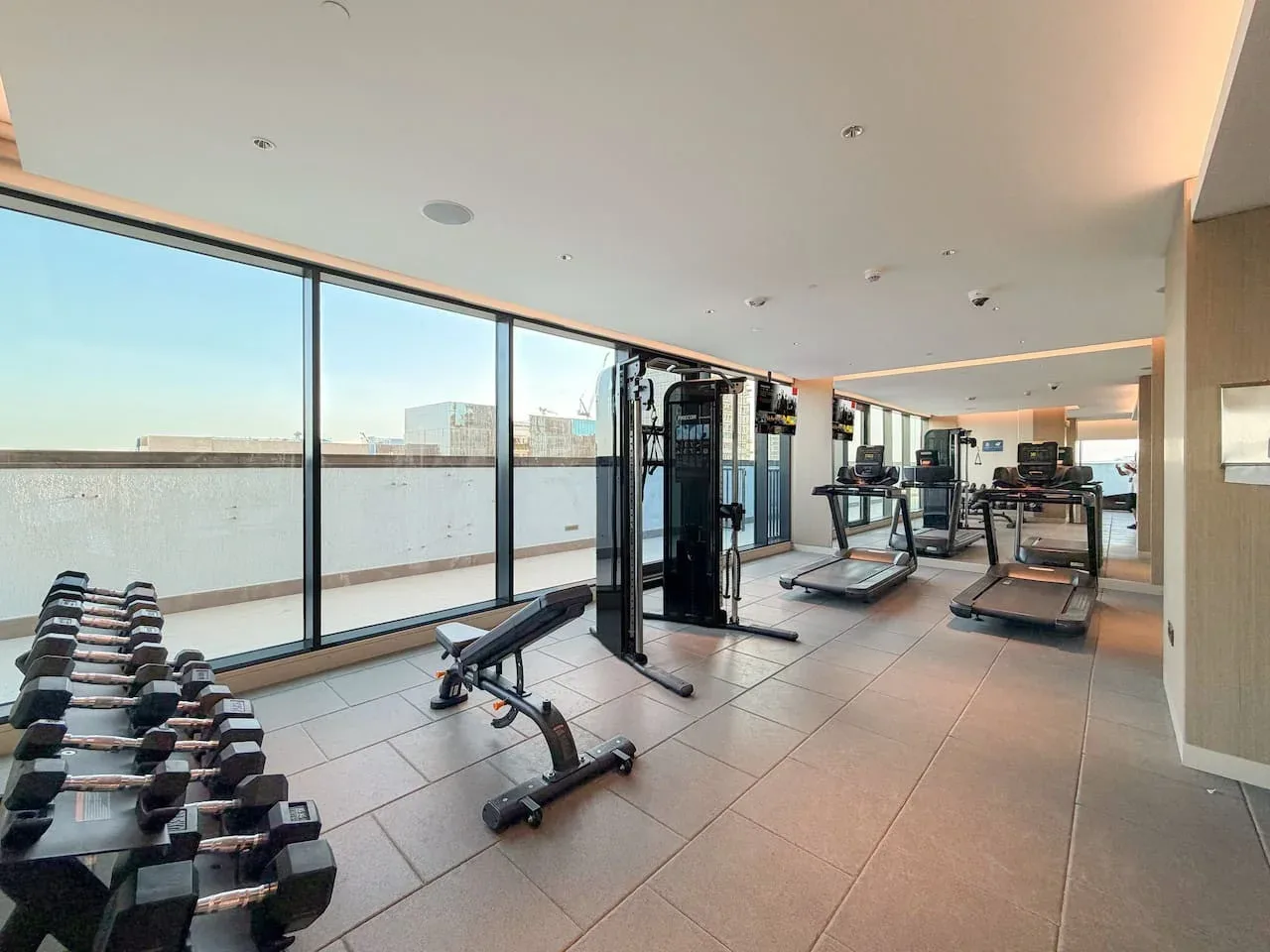 Un gimnasio con máquinas de ejercicio y pesas junto a grandes ventanales.