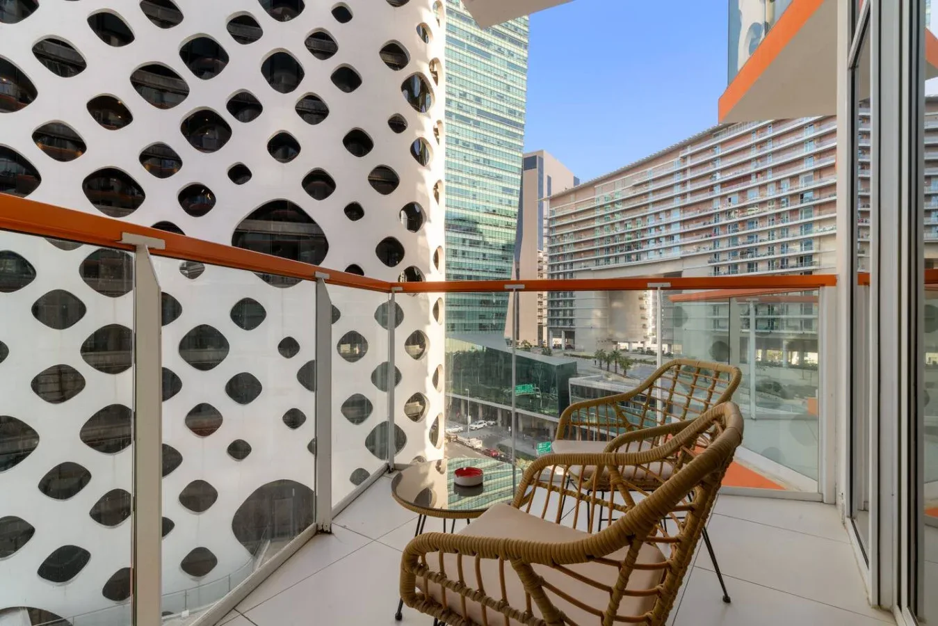 A balcony with woven seating overlooks modern buildings through a glass railing.