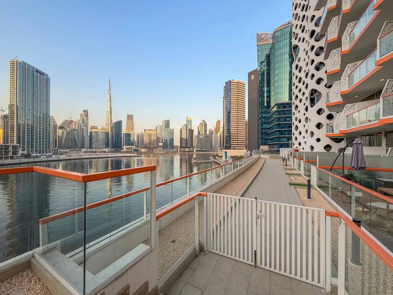 A waterfront terrace overlooks a city skyline, with glass railings along the walkway.