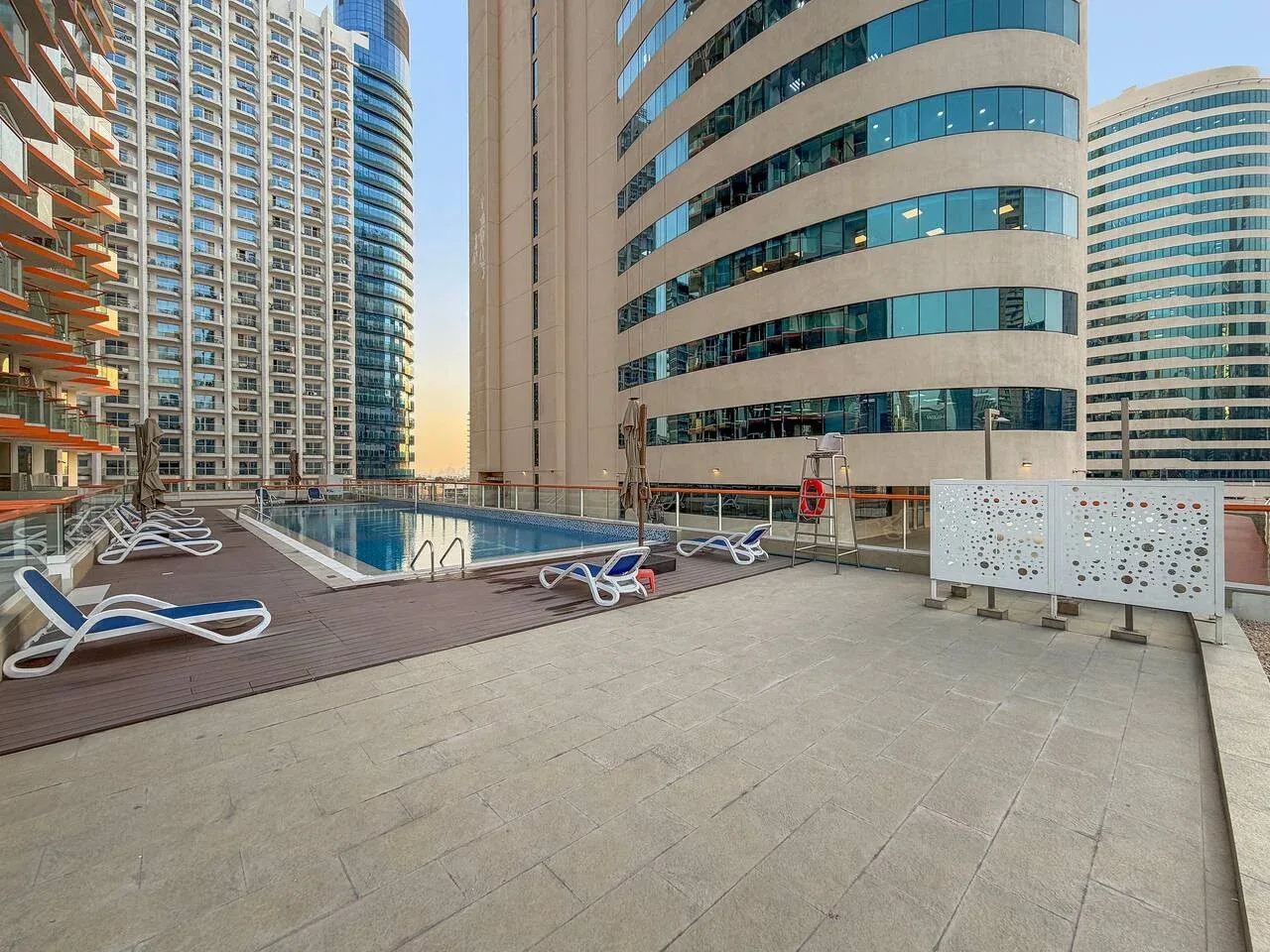 An outdoor pool deck between modern high-rise buildings, with lounge seating nearby.