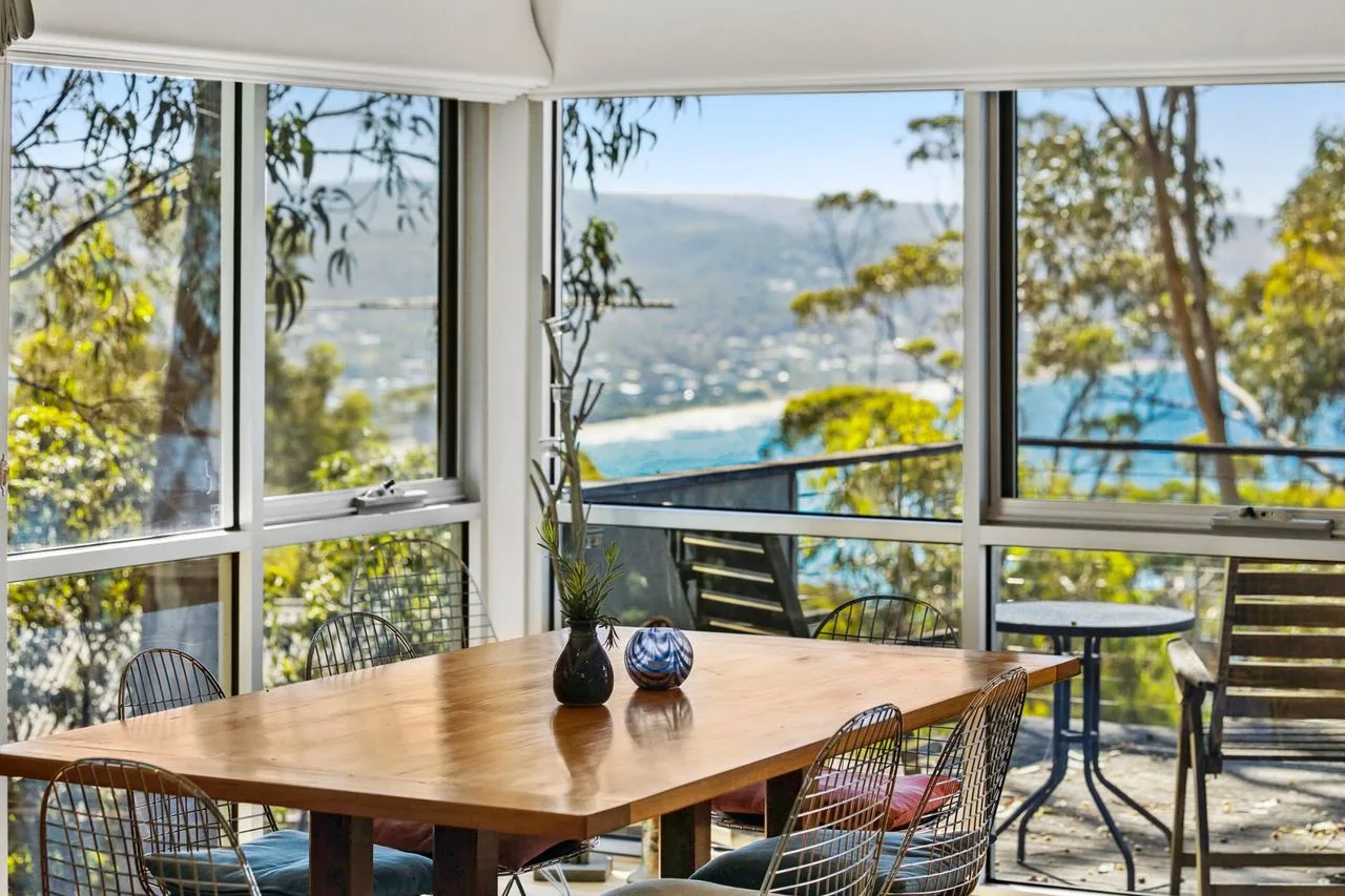 Una mesa de comedor y asientos junto a ventanas con vistas a una colina verde y al agua.