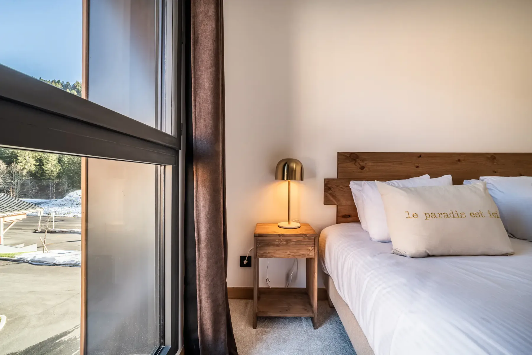 A bedroom with a bed beside a large window and wooden bedside table.
