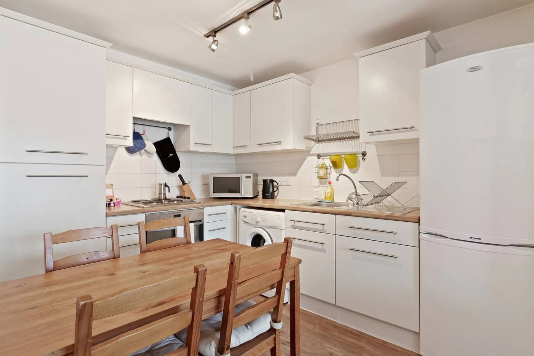 Una cocina luminosa con gabinetes blancos, electrodomésticos y una mesa de comedor de madera.