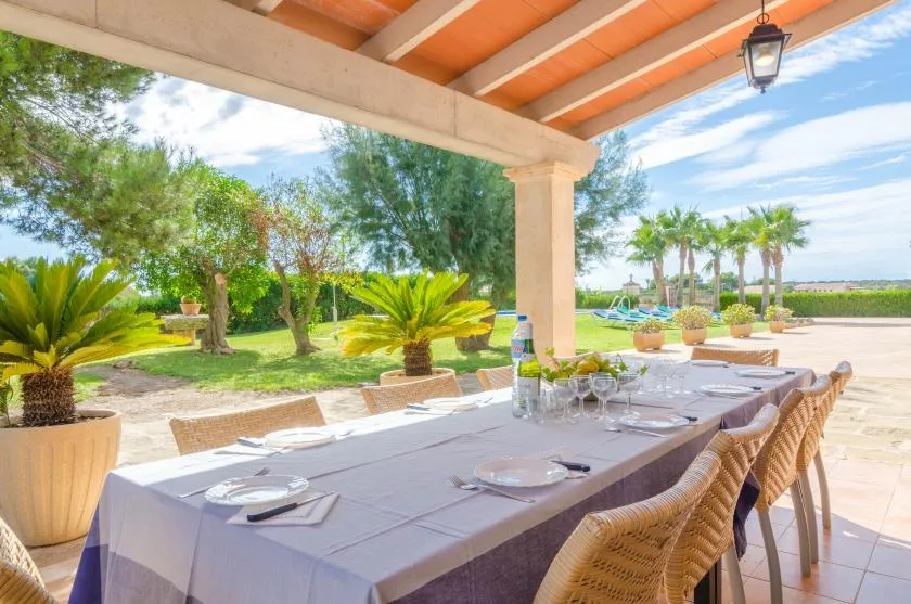 Una zona de comedor exterior cubierta da a un jardín cuidado y una piscina.