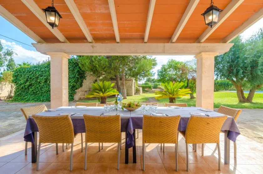 Un comedor exterior cubierto con una mesa larga y asientos tejidos da a un jardín.
