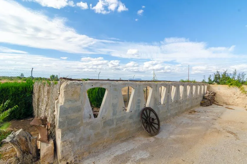 Un muro bajo de hormigón con aberturas inclinadas está junto a un camino de tierra bajo un cielo azul.