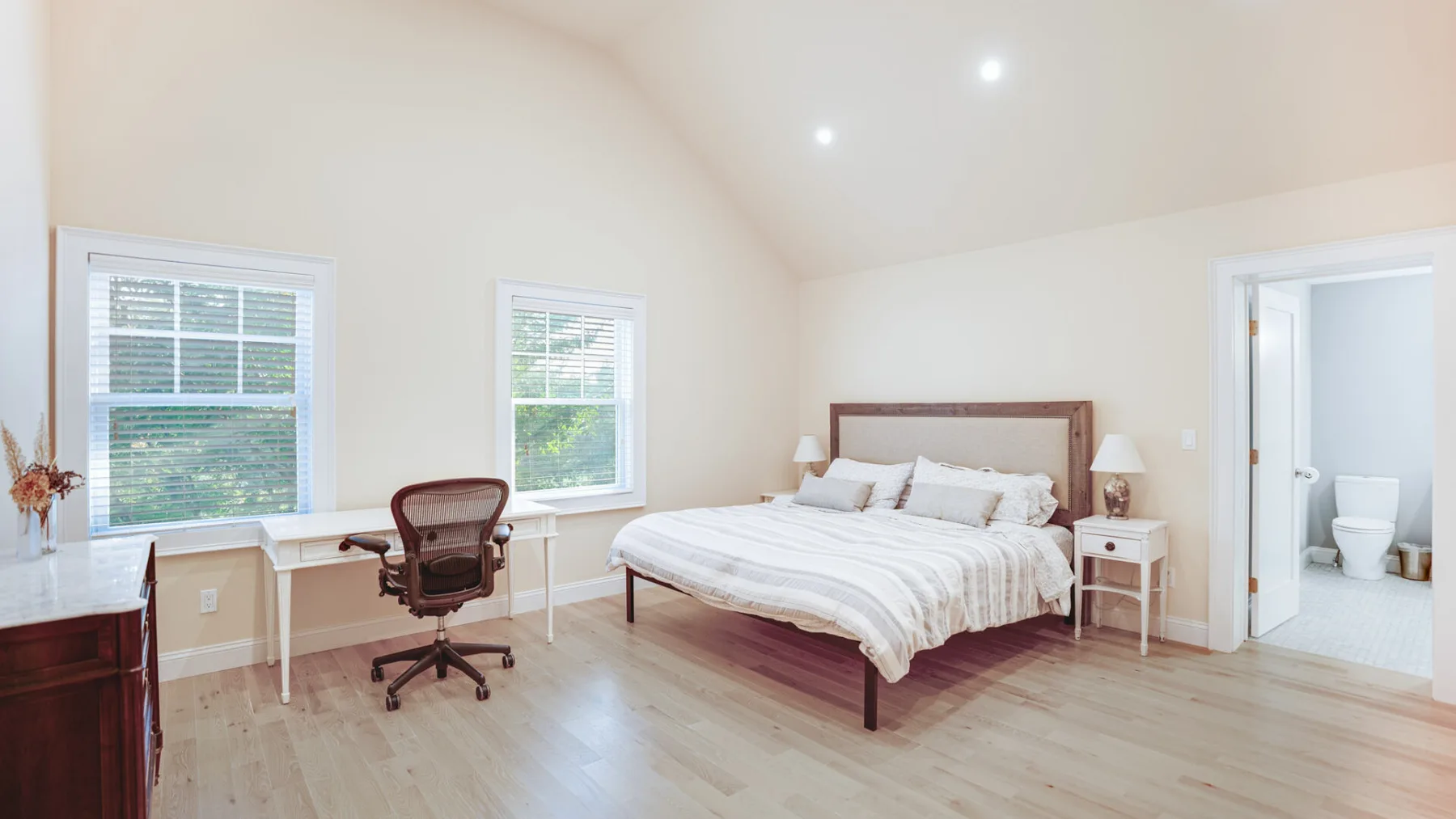 A bright bedroom with a bed and a desk beside the windows.