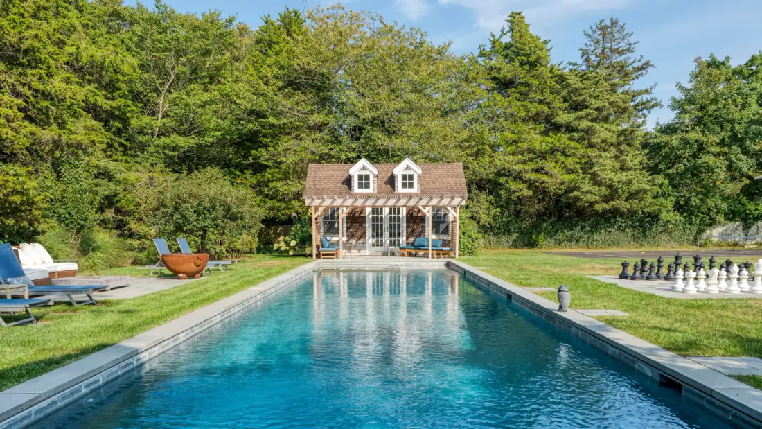 An outdoor swimming pool faces a small pavilion, with trees beyond the lawn.