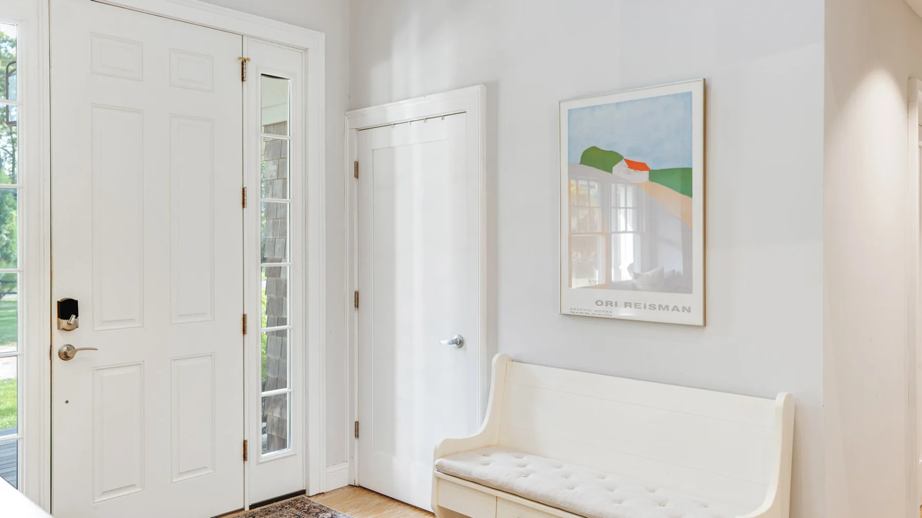 A bright entryway with white doors and a cushioned bench beneath framed artwork.
