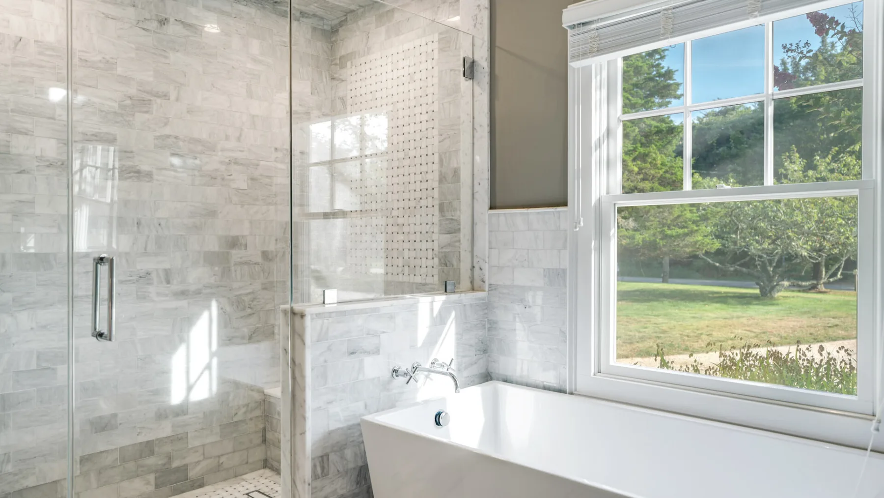 A bright bathroom with a glass shower and a large window overlooking greenery.