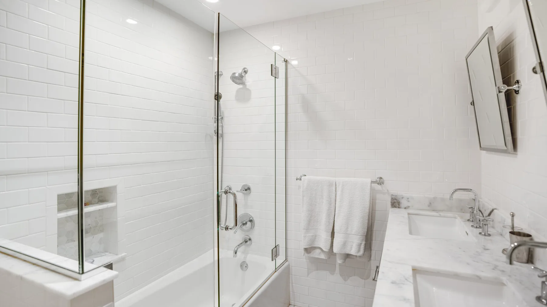 A bright tiled bathroom with a glass shower and a long vanity.