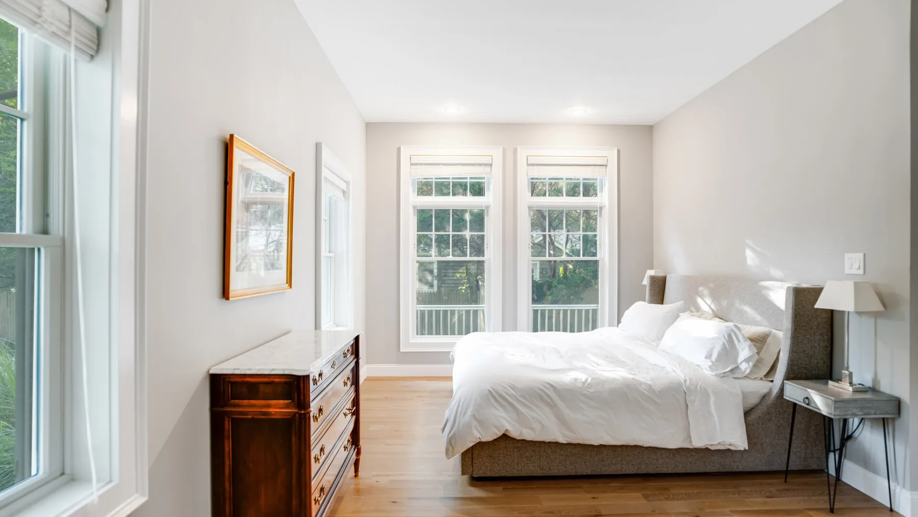 A bright bedroom with a bed beside tall windows and a wooden dresser.