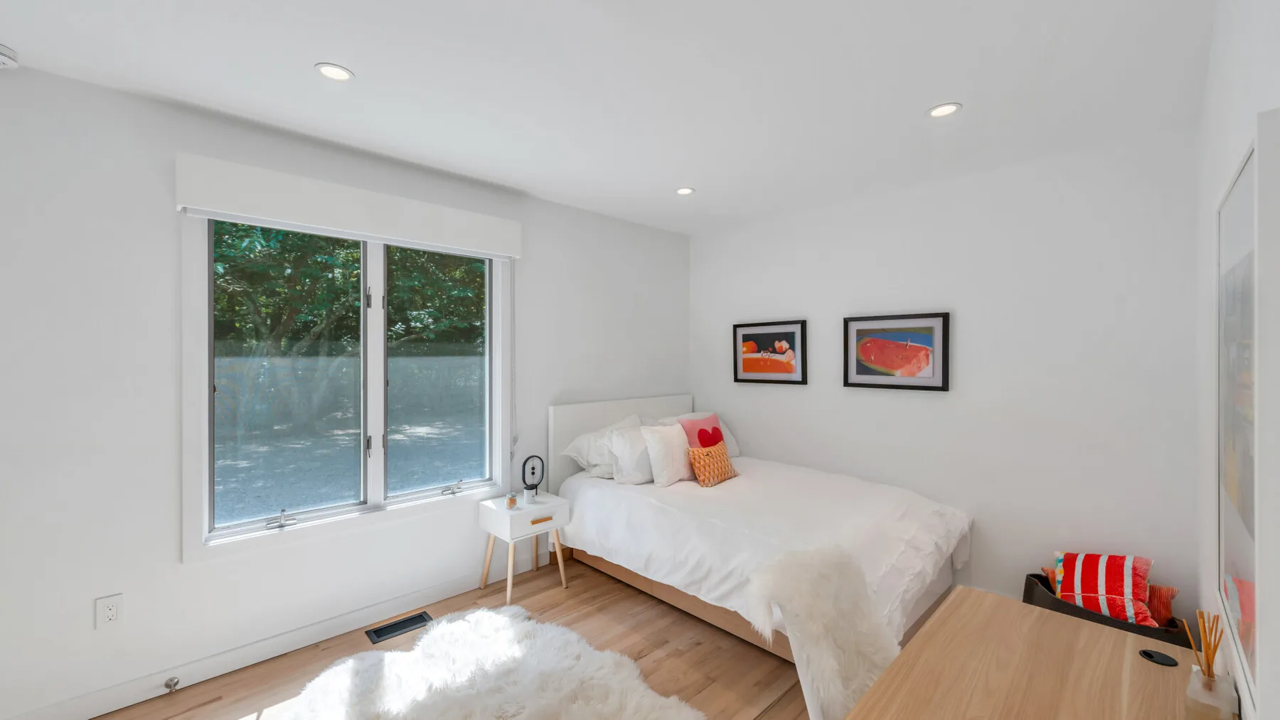 A bright bedroom with a bed beside large windows and a wooden side table.