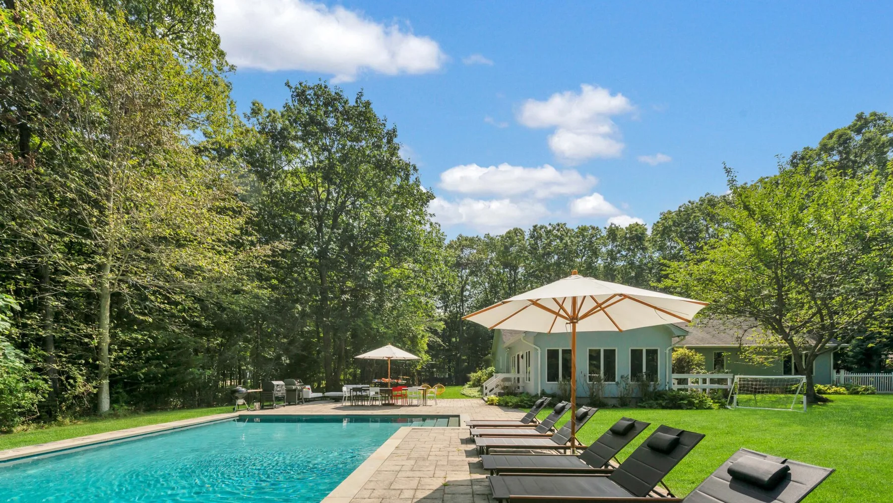 An outdoor swimming pool beside a patio and a house, surrounded by trees.